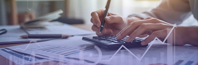 A person calculates financial data on a calculator over reports and charts, symbolizing analysis, planning, and business performance tracking