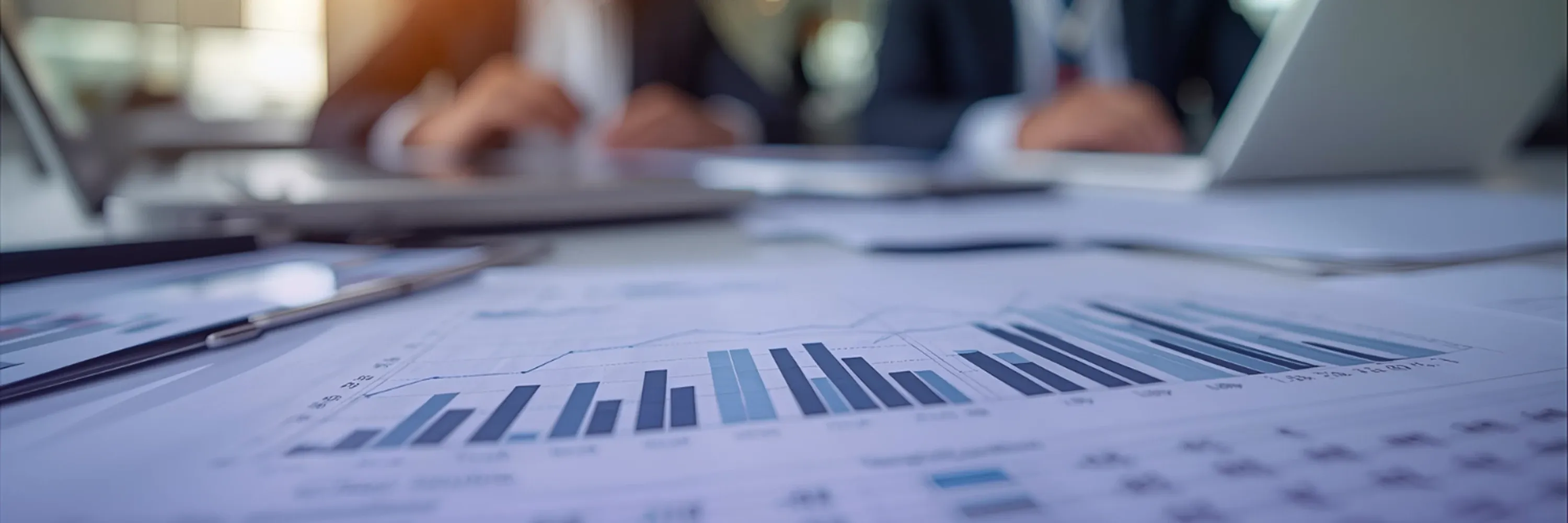 A financial report with bar charts lies on a desk while two business professionals work on laptops in the background