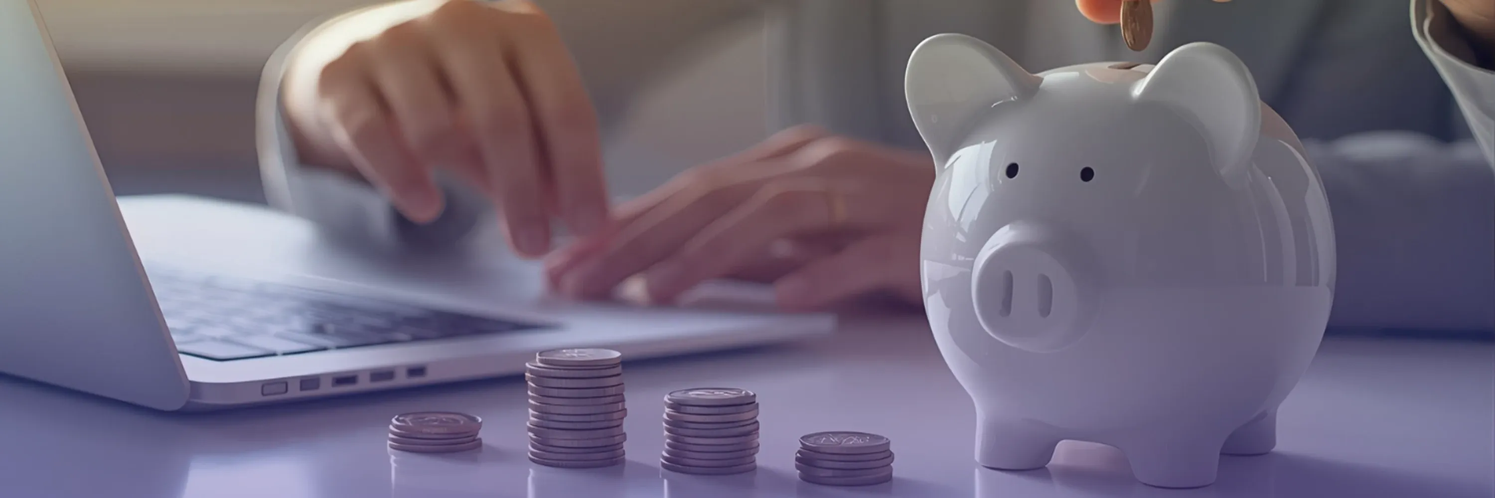 A person places a coin into a piggy bank while working on a laptop, with neatly stacked coins arranged on the desk in front