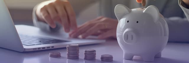 A person places a coin into a piggy bank while working on a laptop, with neatly stacked coins arranged on the desk in front