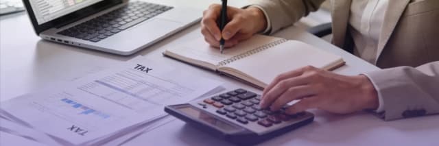 A professional workspace showing hands calculating taxes with a calculator, laptop, notebook, and printed tax documents, illustrating business tax planning and financial analysis