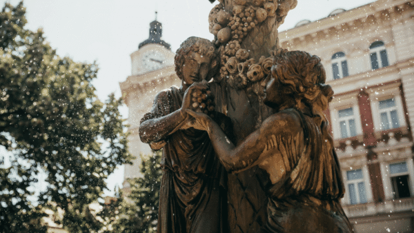 Two women are depicted in a statue, holding hands together in front of a grand building, representing solidarity and connection.