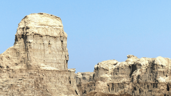 A large, rugged rock formation stands isolated amidst the endless sands of a sunlit desert.